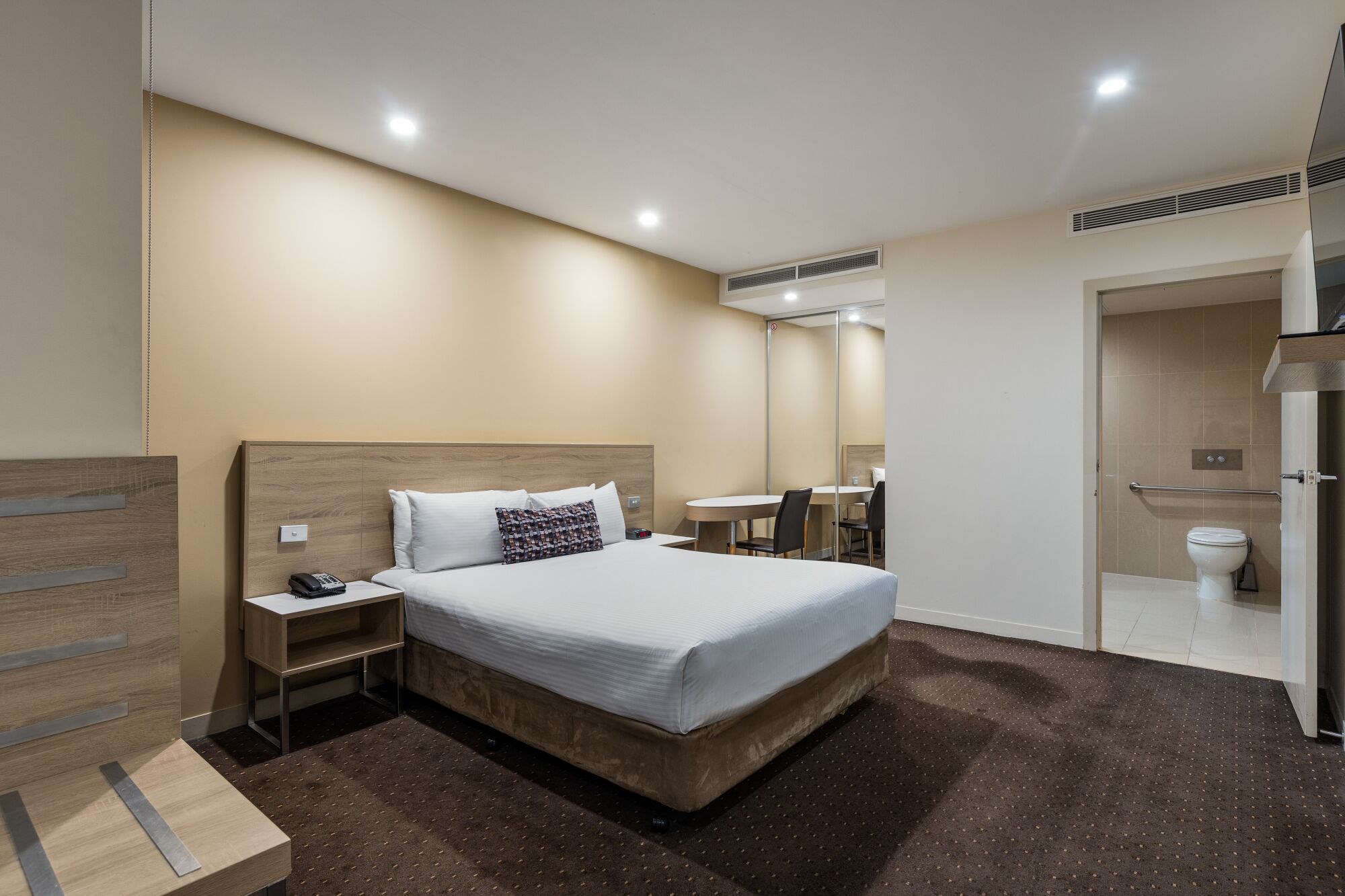 Een hotelkamer met een bed, nachtkastje en een klein bureau met stoel. Een badkamer met toilet en wastafel is zichtbaar door een geopende deur.