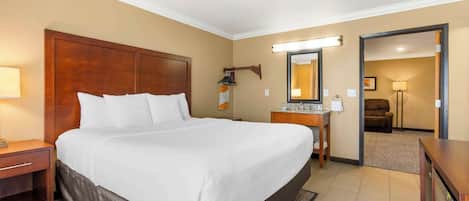 Habitación de hotel con una cama grande, cabecera de madera, mesita de noche, espejo, escritorio con lámpara y vista a otra habitación.