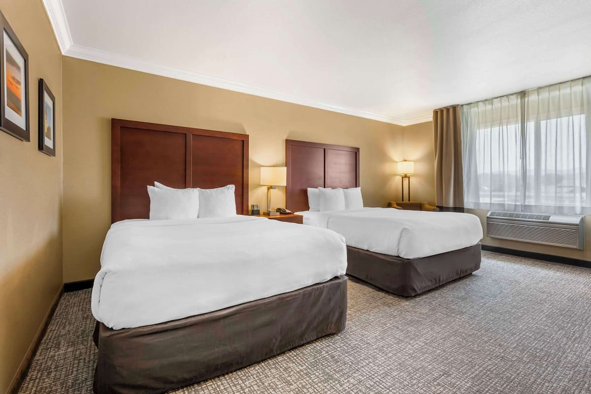 A hotel room with two beds, a large window with curtains, a wall-mounted air conditioner, and framed pictures on the wall.