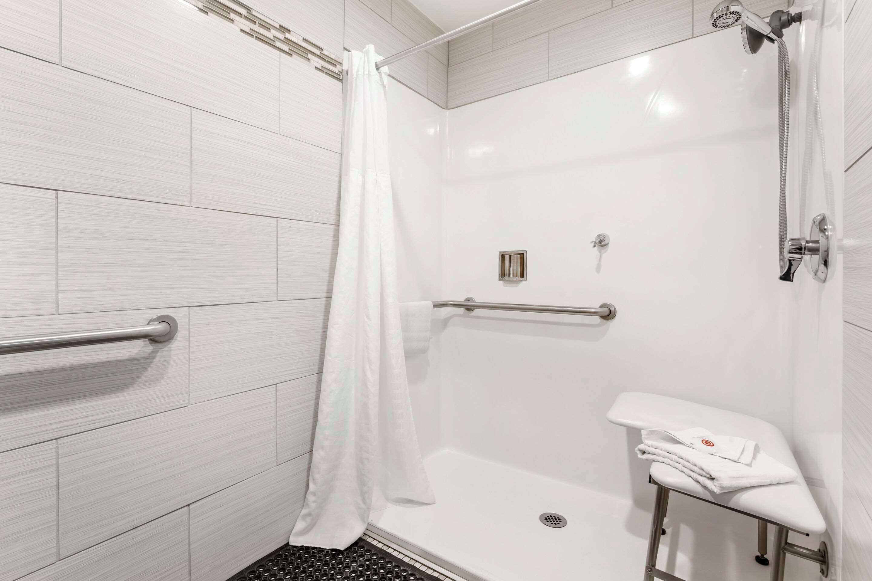 A modern bathroom with white tiles, a shower with a handheld showerhead, a grab bar, and a small bench with towels.