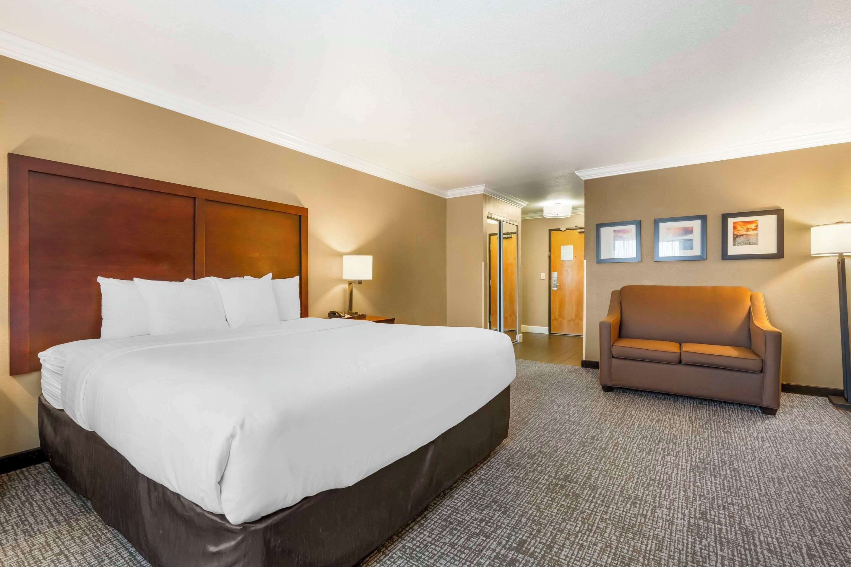 A hotel room with a large bed, a brown armchair, a desk with a lamp, and framed pictures on the wall.