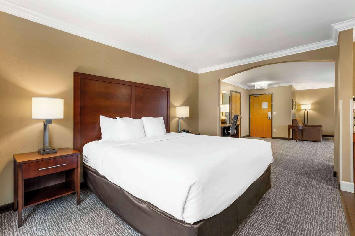 A hotel room with a large bed, a nightstand with a lamp, a desk with a chair, and a mirror.