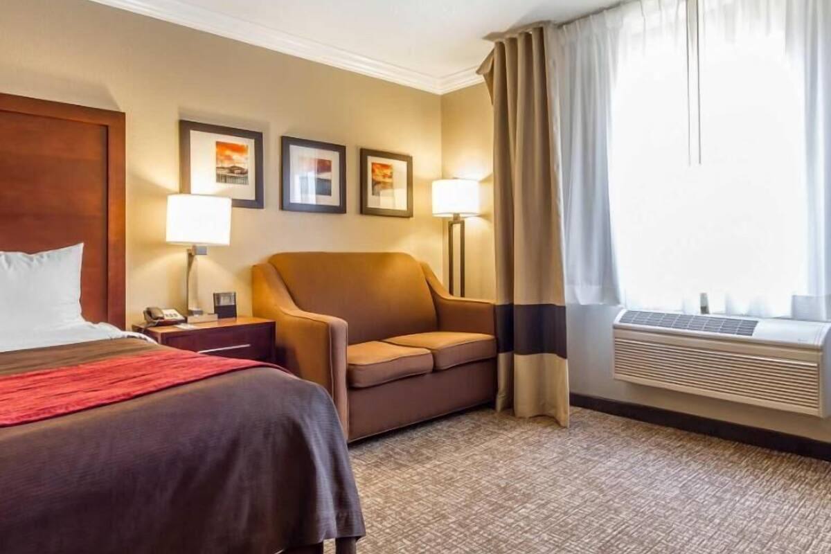 A hotel room with a bed, a sofa, a desk with a lamp, framed pictures on the wall, and an air conditioning unit.