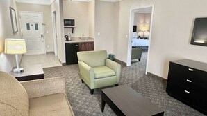 Une chambre d  ’hôtel avec un canapé, un fauteuil, une table basse et un téléviseur. Elle dispose d’une kitchenette équipée d’un four à micro-ondes et d’un évier. La chambre est dotée d’un revêtement de sol en moquette et d’une porte donnant sur une autre pièce.