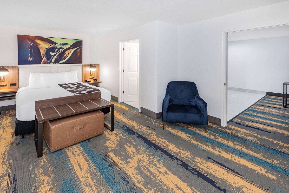 A hotel room with a bed, bedside table, lamp, chair, and a large painting on the wall.