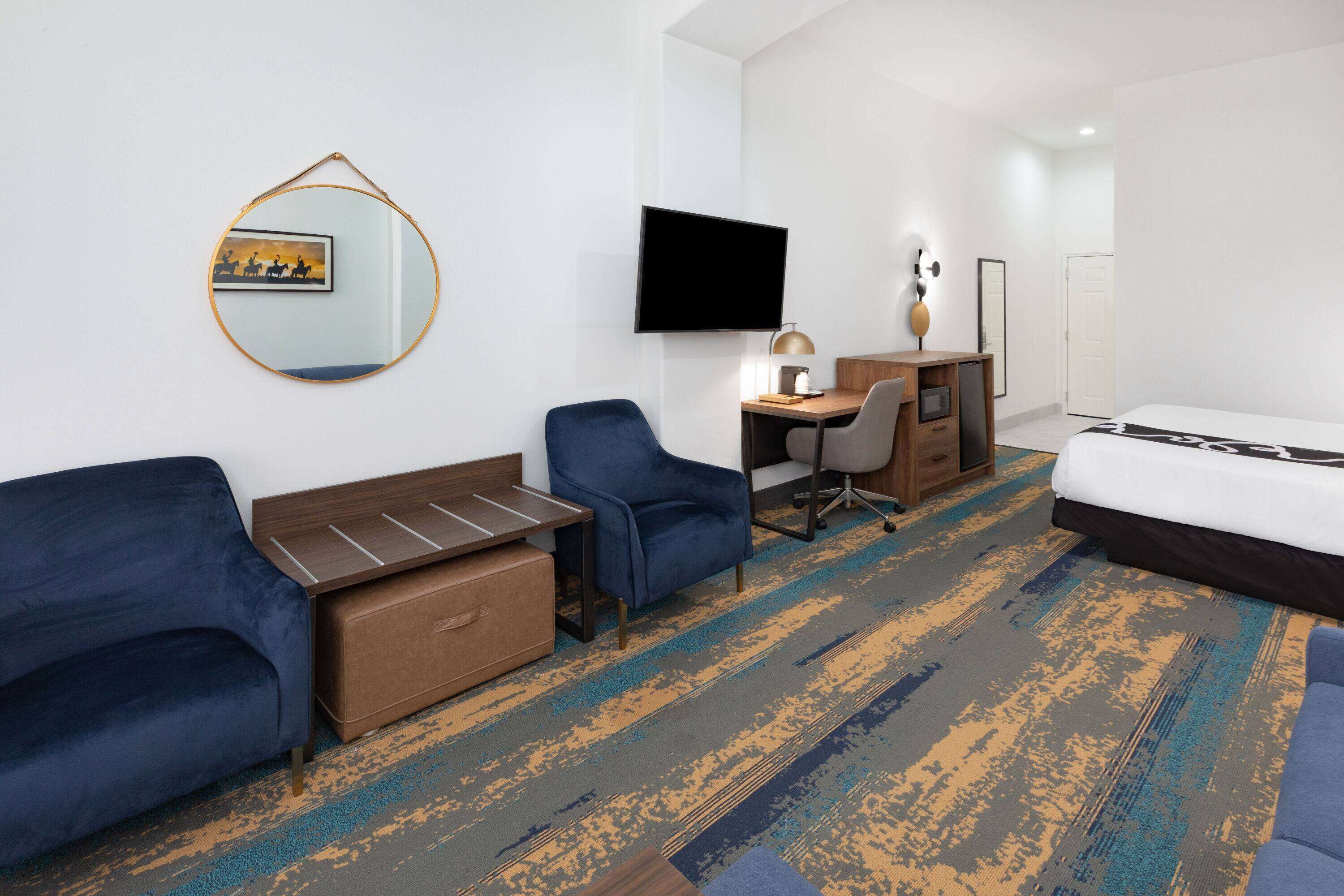 A hotel room with a bed, a desk with a chair, a sofa, a mirror, and a flat-screen TV mounted on the wall.