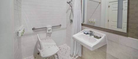 A bathroom with a white sink, a mirror, a shower with a handheld showerhead, a grab bar, and a white chair.