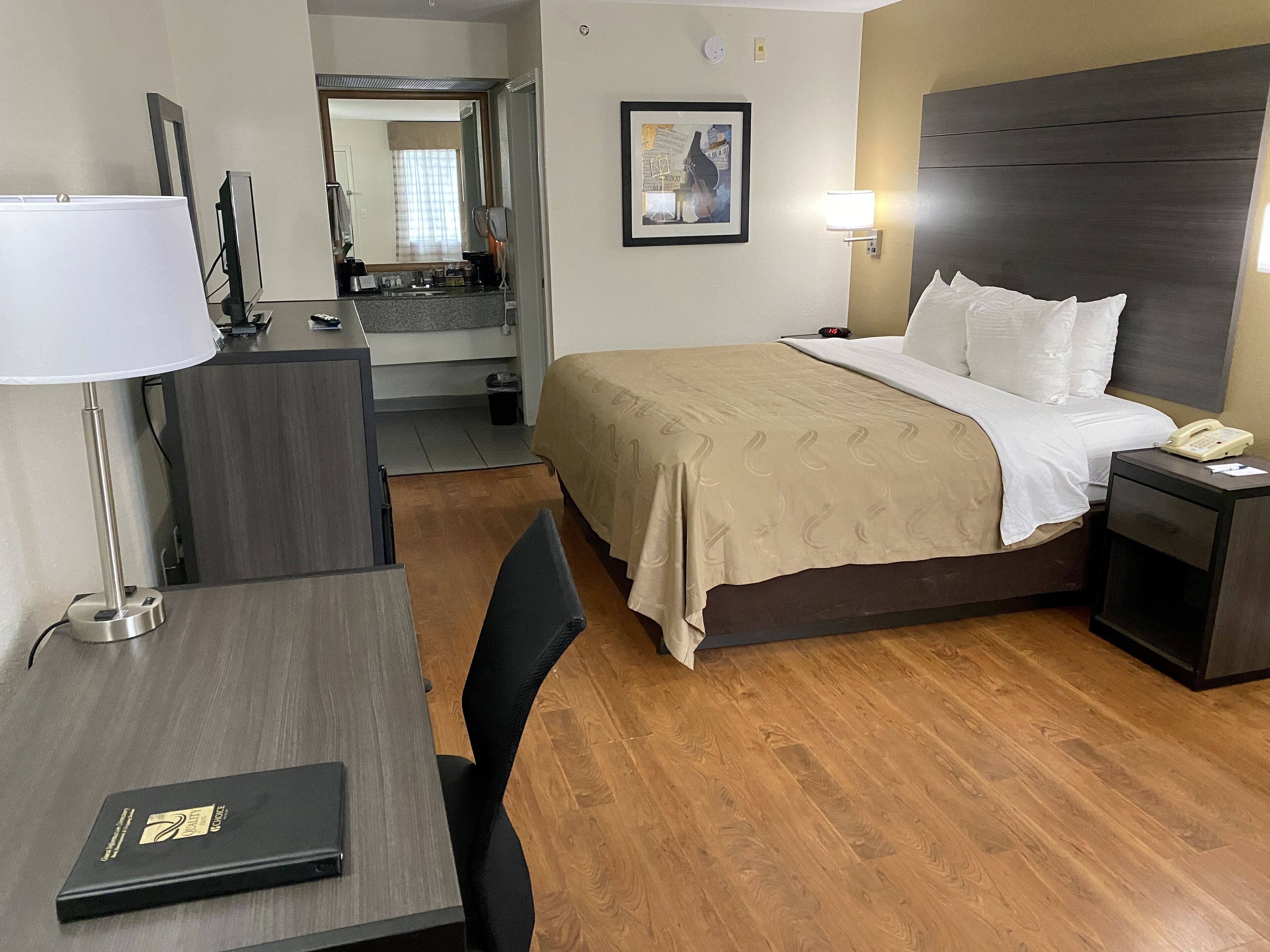 Une chambre d’hôtel comprenant un lit, une table de chevet, une lampe, un bureau, une chaise et une salle de bain visible à travers une porte ouverte.