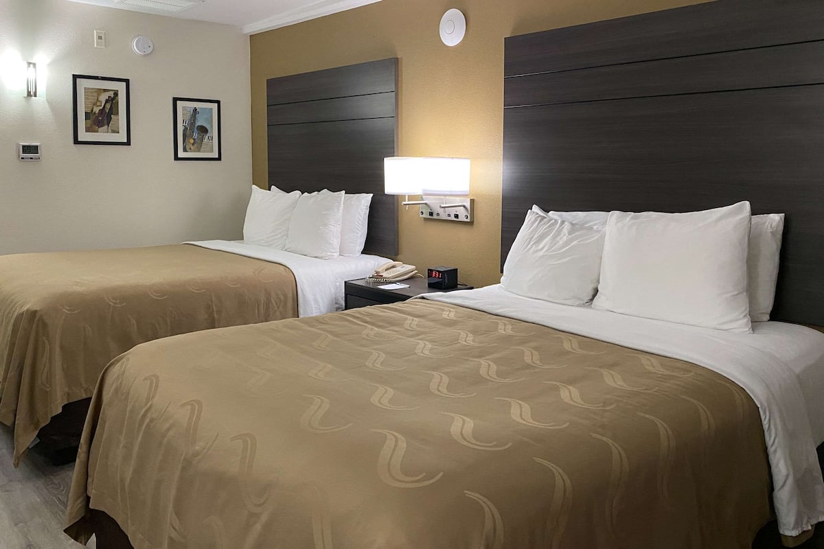 A hotel room with two beds, each with a beige bedspread and white pillows, a bedside table with a lamp, and framed artwork on the wall.