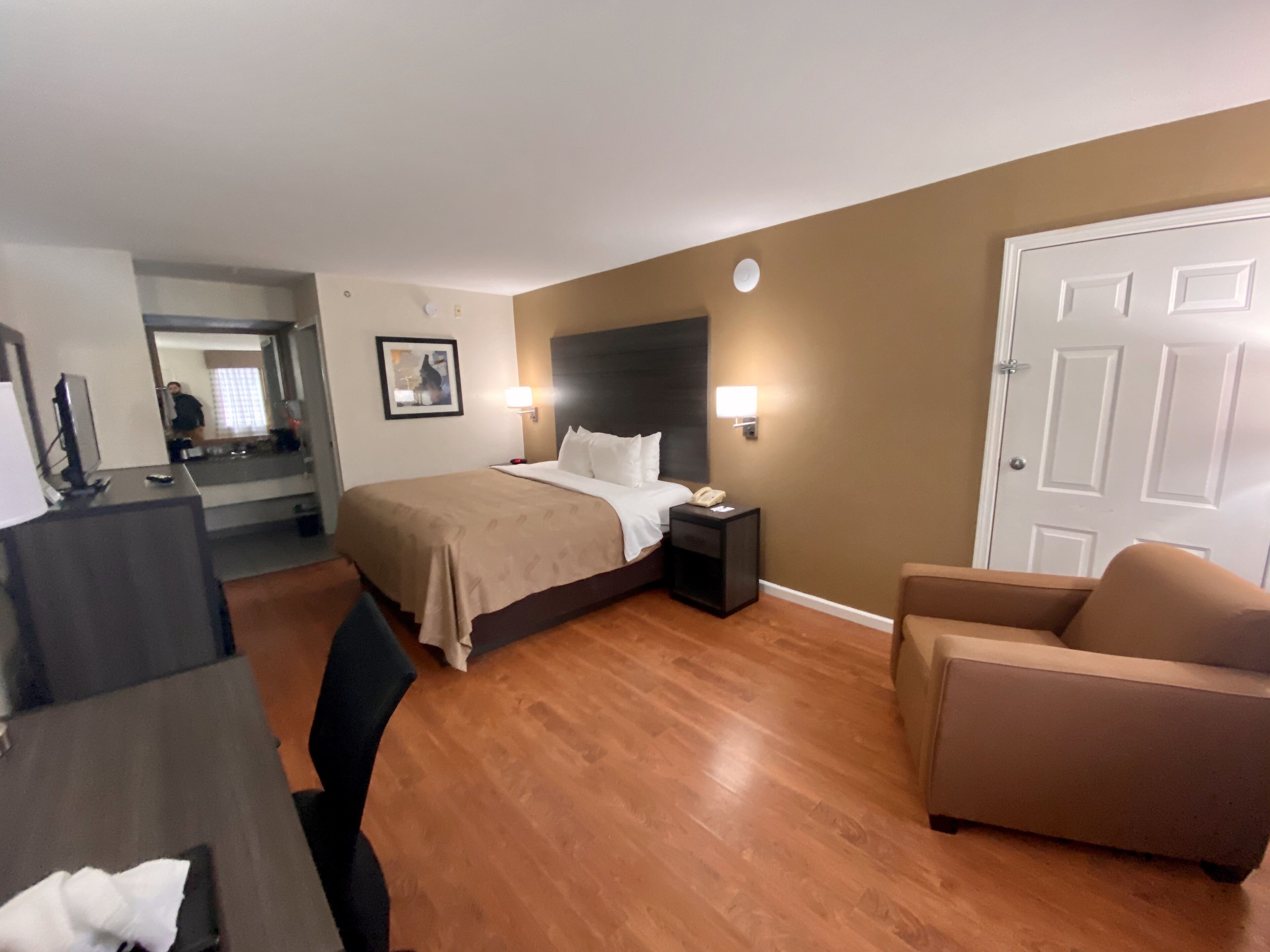 A hotel room with a bed, bedside table, lamp, chair, desk, and a door.