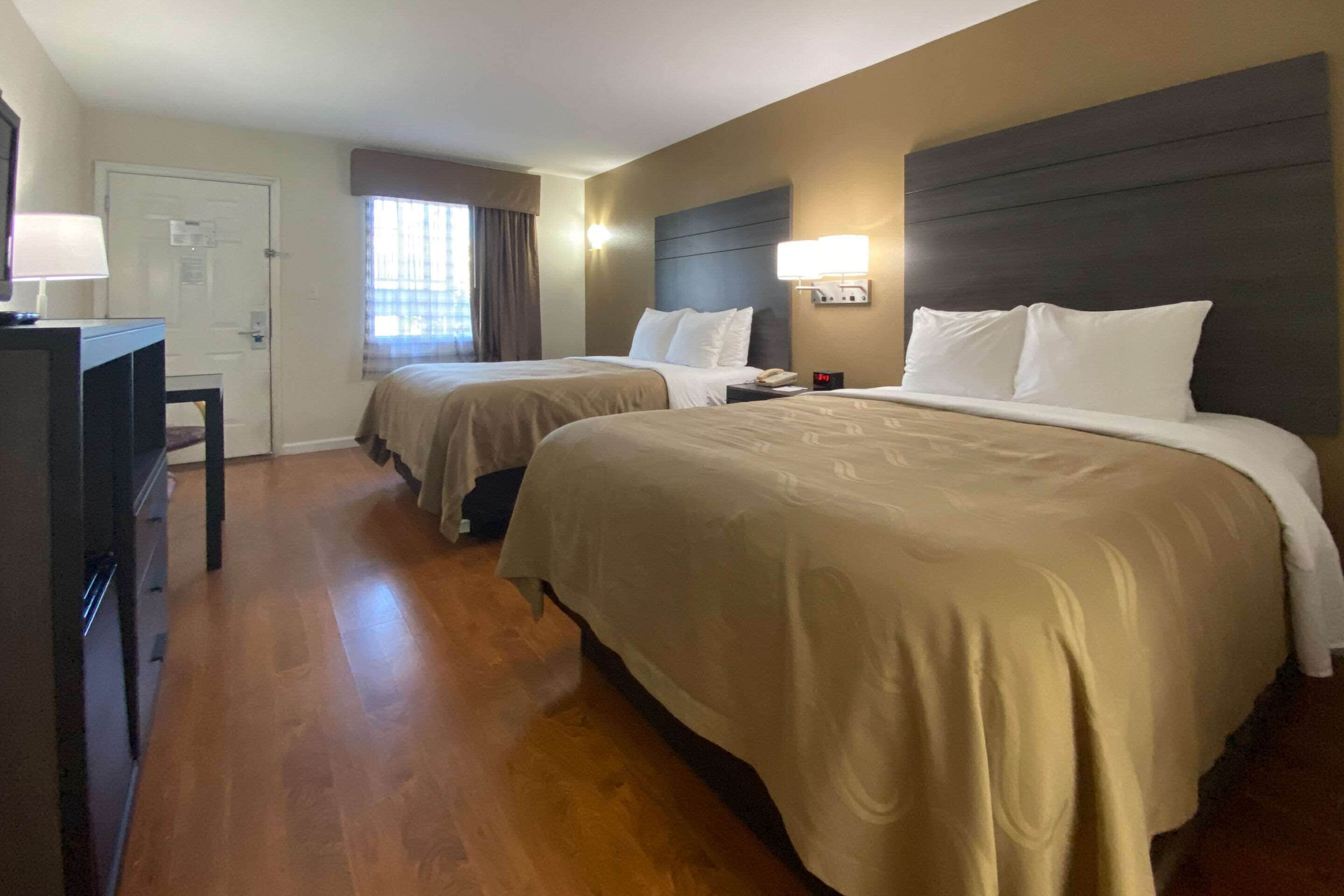 A hotel room with two beds, a desk, a lamp, and a window with curtains.