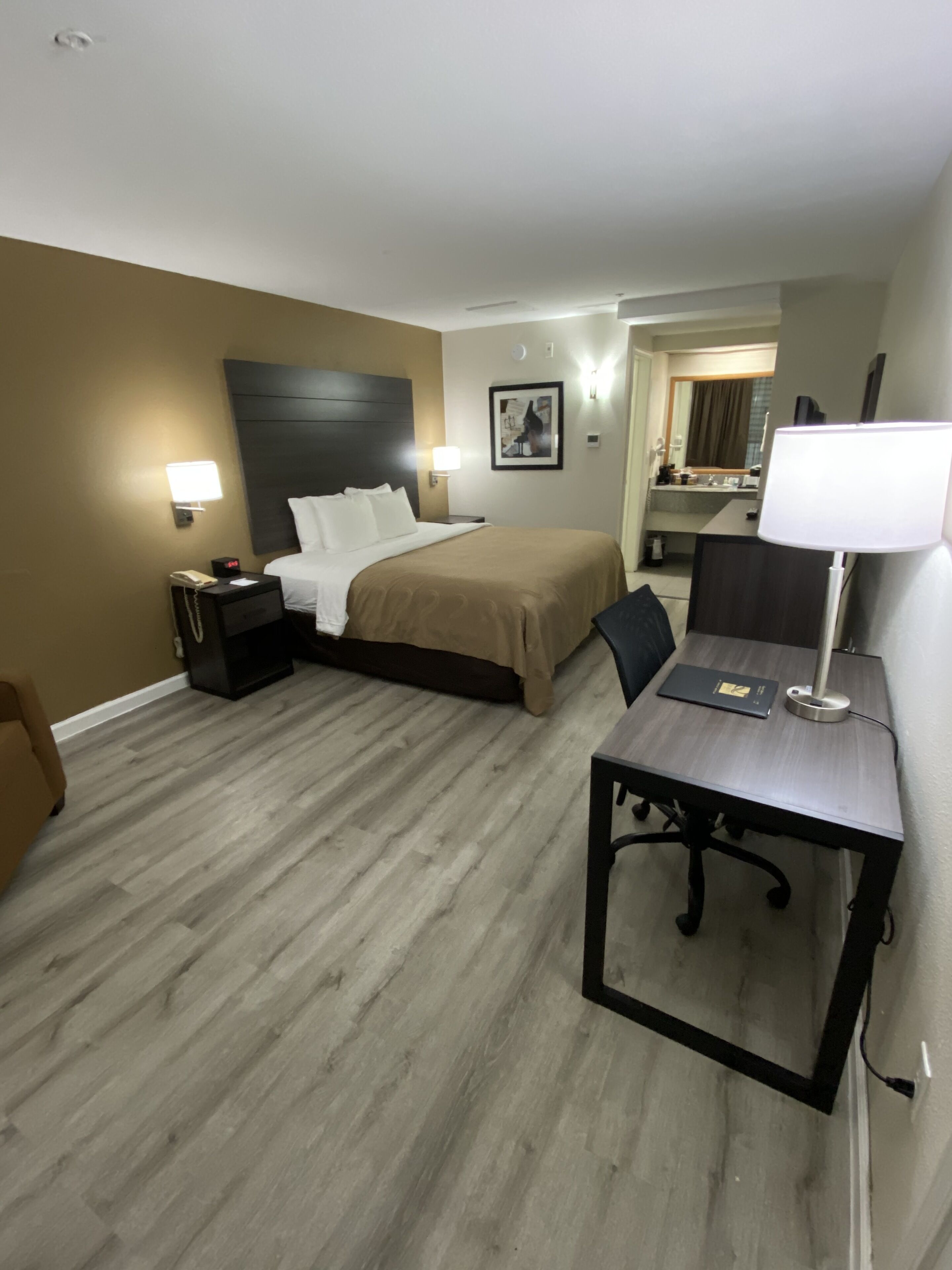 Quarto de hotel com cama, mesas de cabeceira, escrivaninha e cadeira, e um banheiro visível ao fundo.