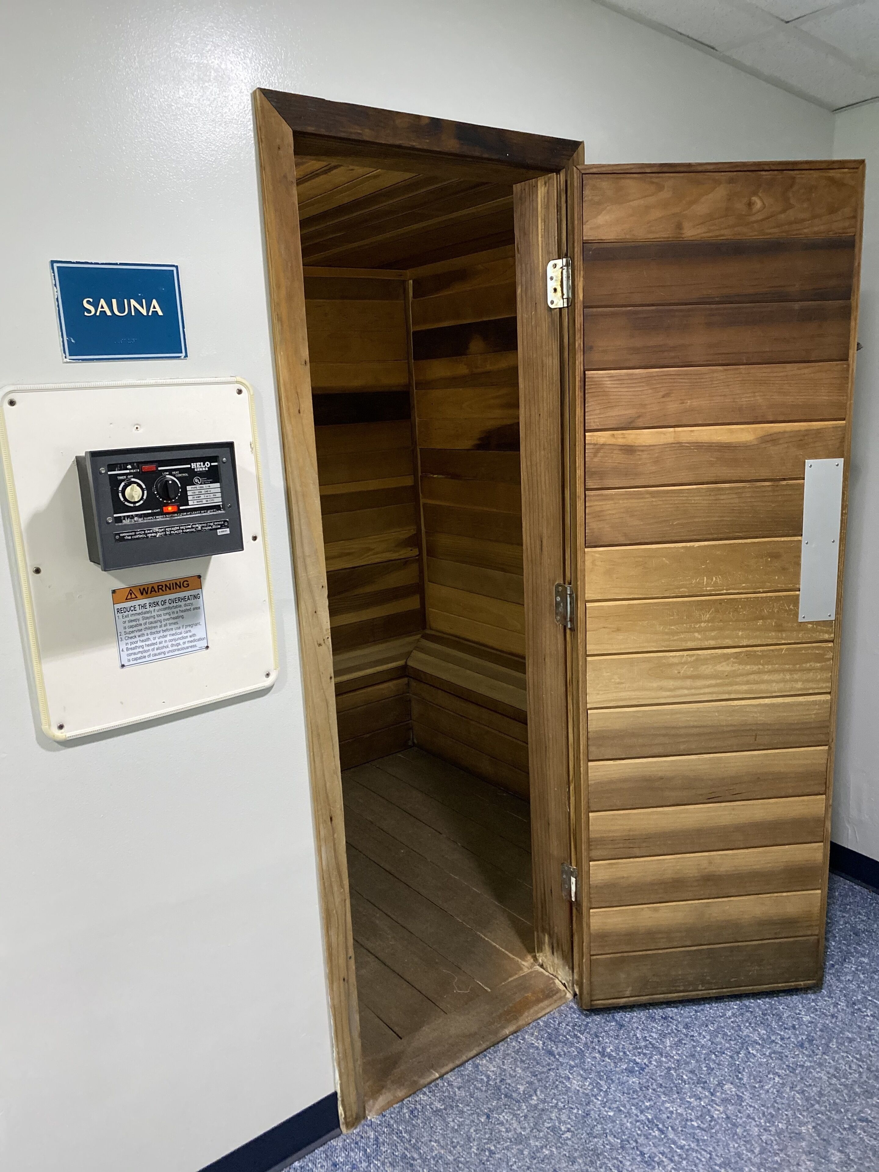 Un cuarto de sauna con paredes de madera y una puerta, un panel de control con un letrero de advertencia y un cartel que indica el propósito de la sala.