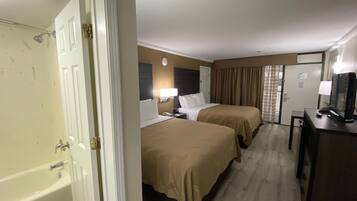A hotel room with two beds, a desk with a computer, a TV, a bathroom with a shower, and a clock on the wall.