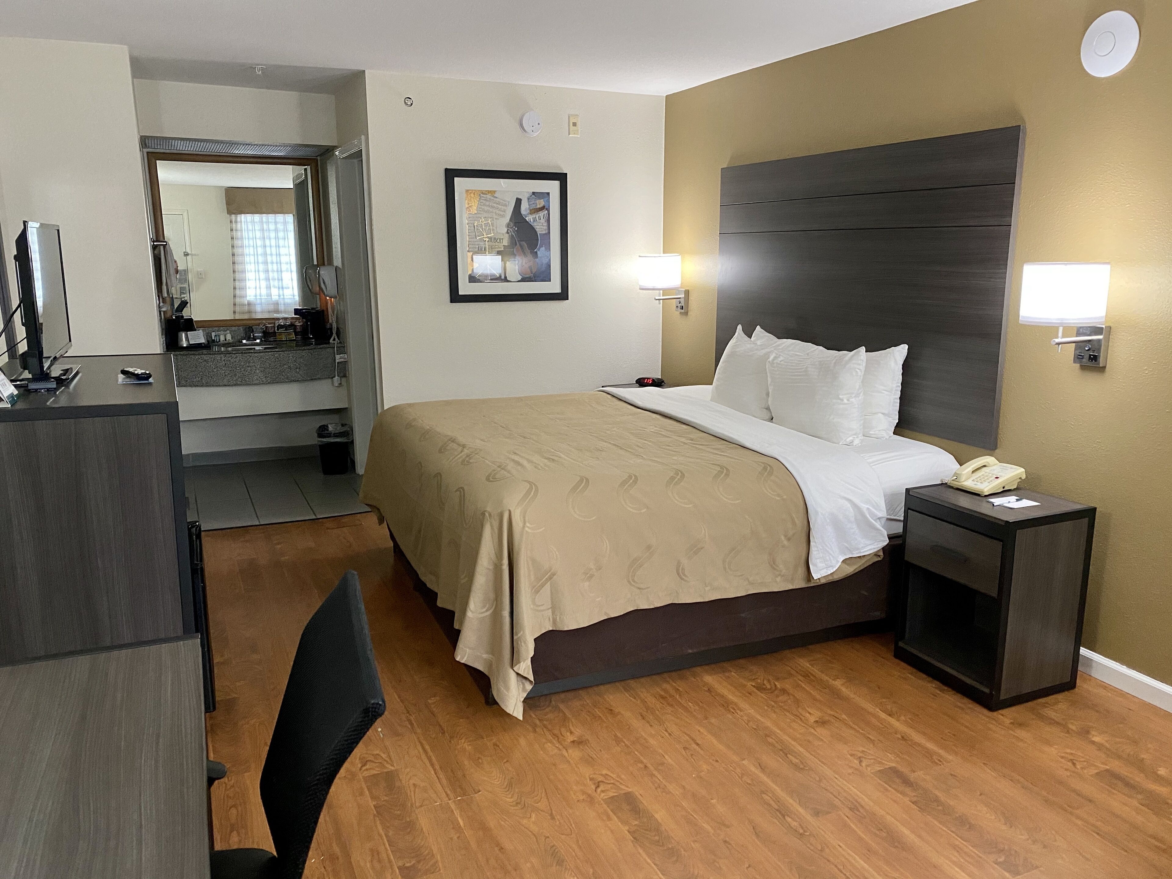 A hotel room with a bed, bedside table, desk, chair, and a bathroom visible through an open door.