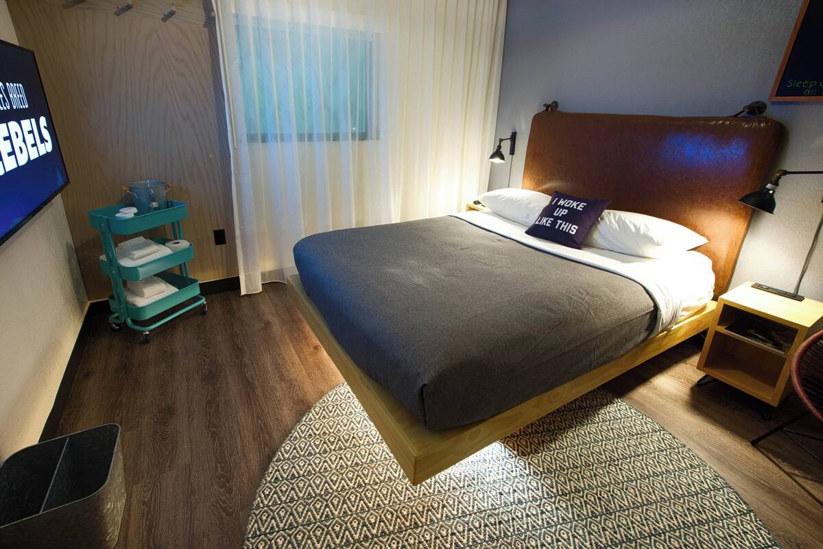 Une chambre d’hôtel moderne avec un grand lit, une tête de lit en bois, une petite table de chevet avec une lampe, une table d’appoint de couleur turquoise avec un panier, et un téléviseur fixé au mur.
