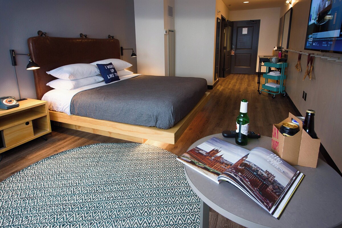 A modern hotel room with a bed, bedside table, lamp, magazine, and a small cart with bottles.