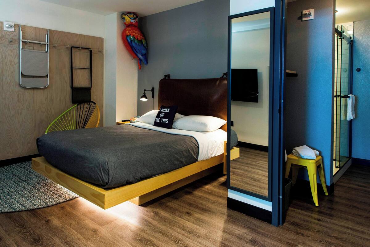 A modern bedroom with a wooden bed, a small stool, a chair, and a colorful bird painting.