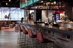 A modern bar area with a long counter, multiple stools, and a variety of bottles and glasses. - Moxy New Orleans Downtown French Quarter Area (New Orleans)