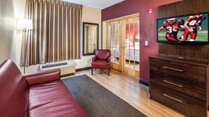 A hotel room with a red sofa, a wooden dresser, a television displaying a football game, a red chair, a window with curtains, and a radiator.