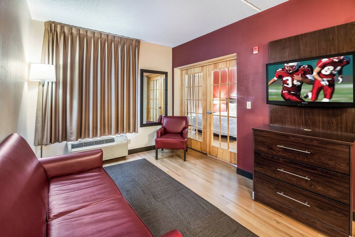 Ett hotellrum med en röd soffa, en byrå i trä, en tv som visar en fotbollsmatch, en röd stol, ett fönster med gardiner och ett element.