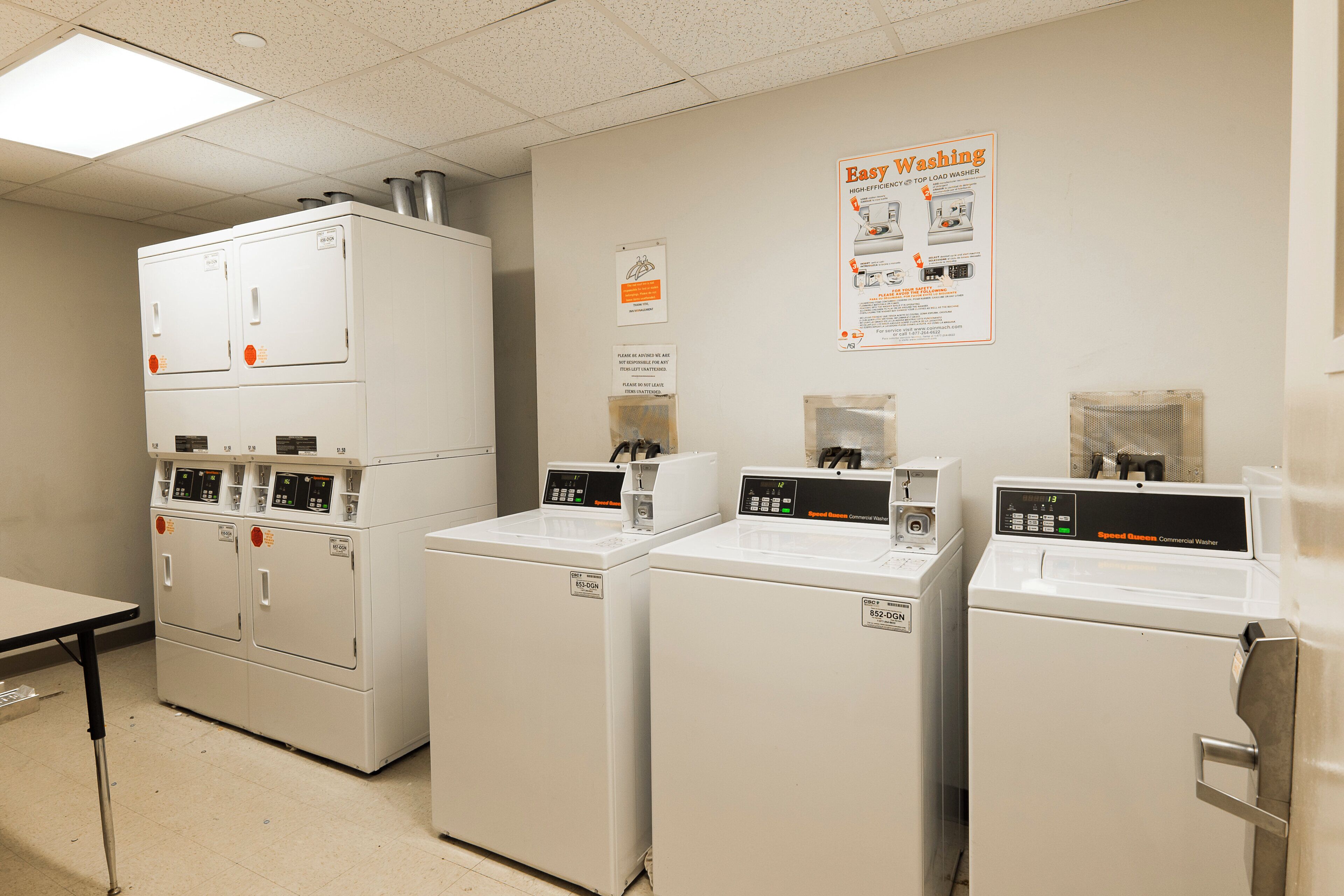A laundry facility with multiple washing machines and dryers, a poster on the wall, and a table.