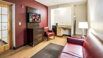 A compact living space with a flat-screen TV mounted on the wall, a wooden cabinet, a red leather sofa, and a kitchenette area.