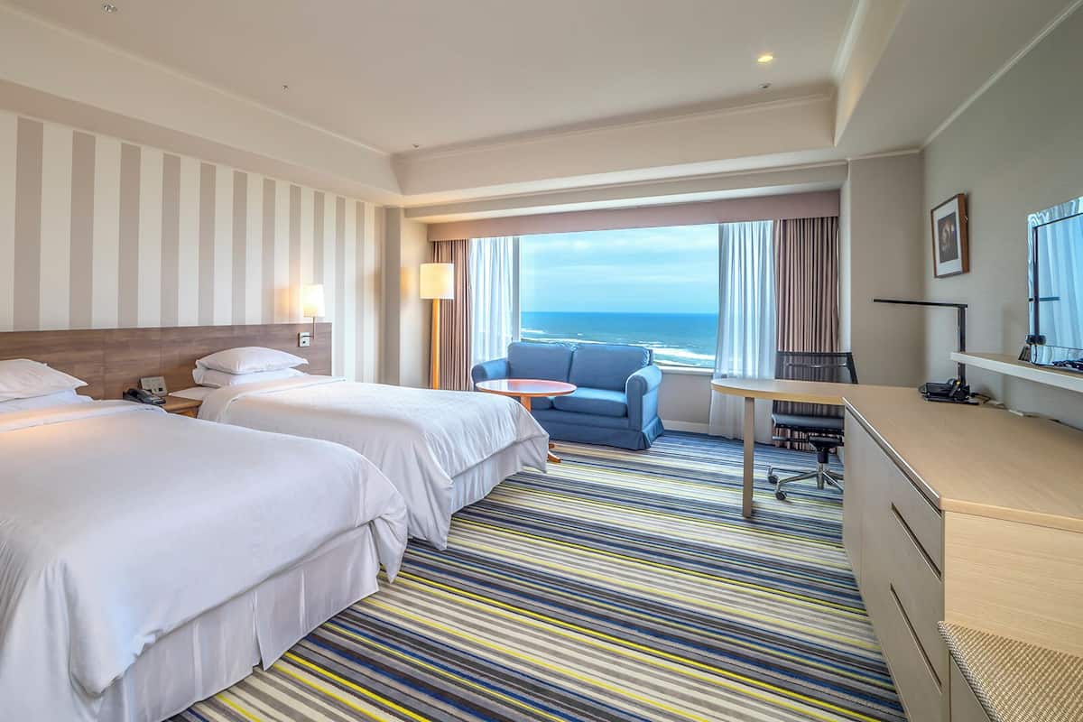A hotel room with two beds, a desk, a chair, and a view of the ocean.