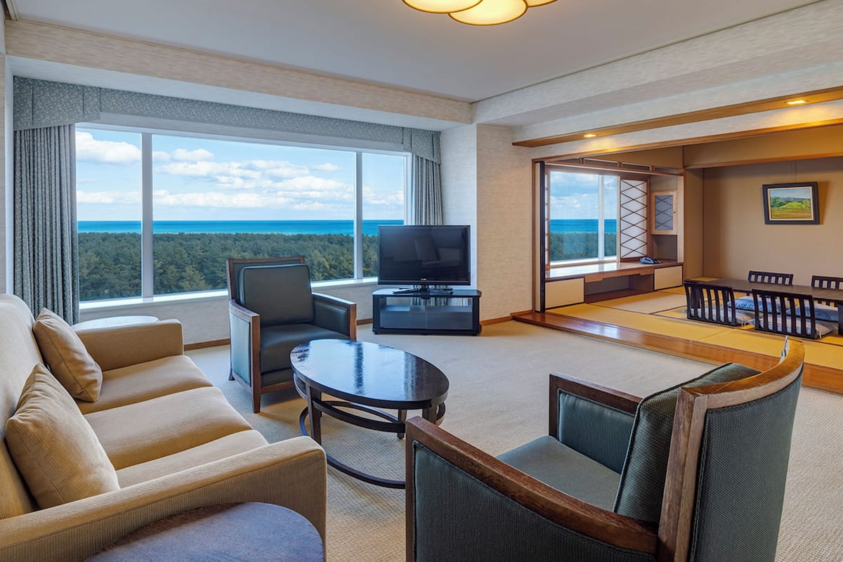 A hotel room with a sofa, armchair, TV, and a view of the ocean.