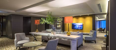 A modern hotel lobby with a central seating area, a flat-screen TV, and a large plant.