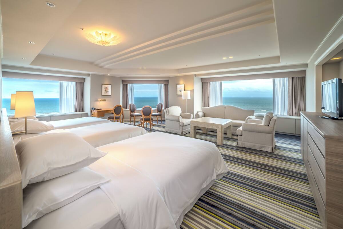 A hotel room with two beds, a dining area, a TV, and a view of the sea.