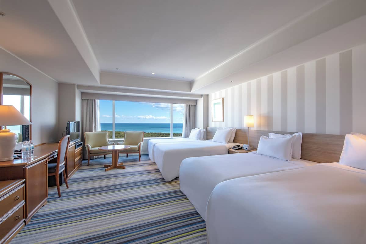 A hotel room with two beds, a desk, a chair, a TV, and a large window with a view of the ocean.