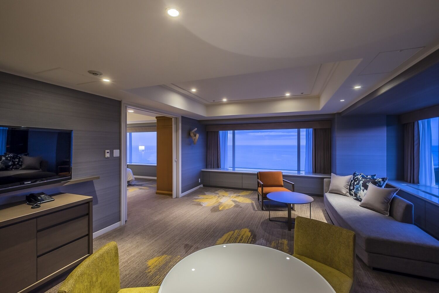 A modern hotel room with a large bed, a TV, a desk with a chair, a sofa, and a window with a view of the ocean.