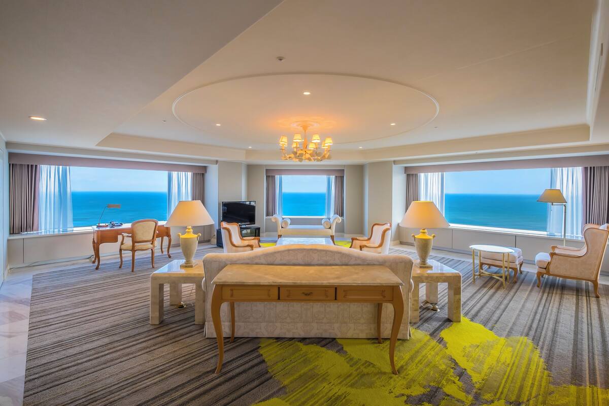 A spacious hotel room with a large bed, two armchairs, a desk, and a TV. The room has large windows with a view of the ocean.