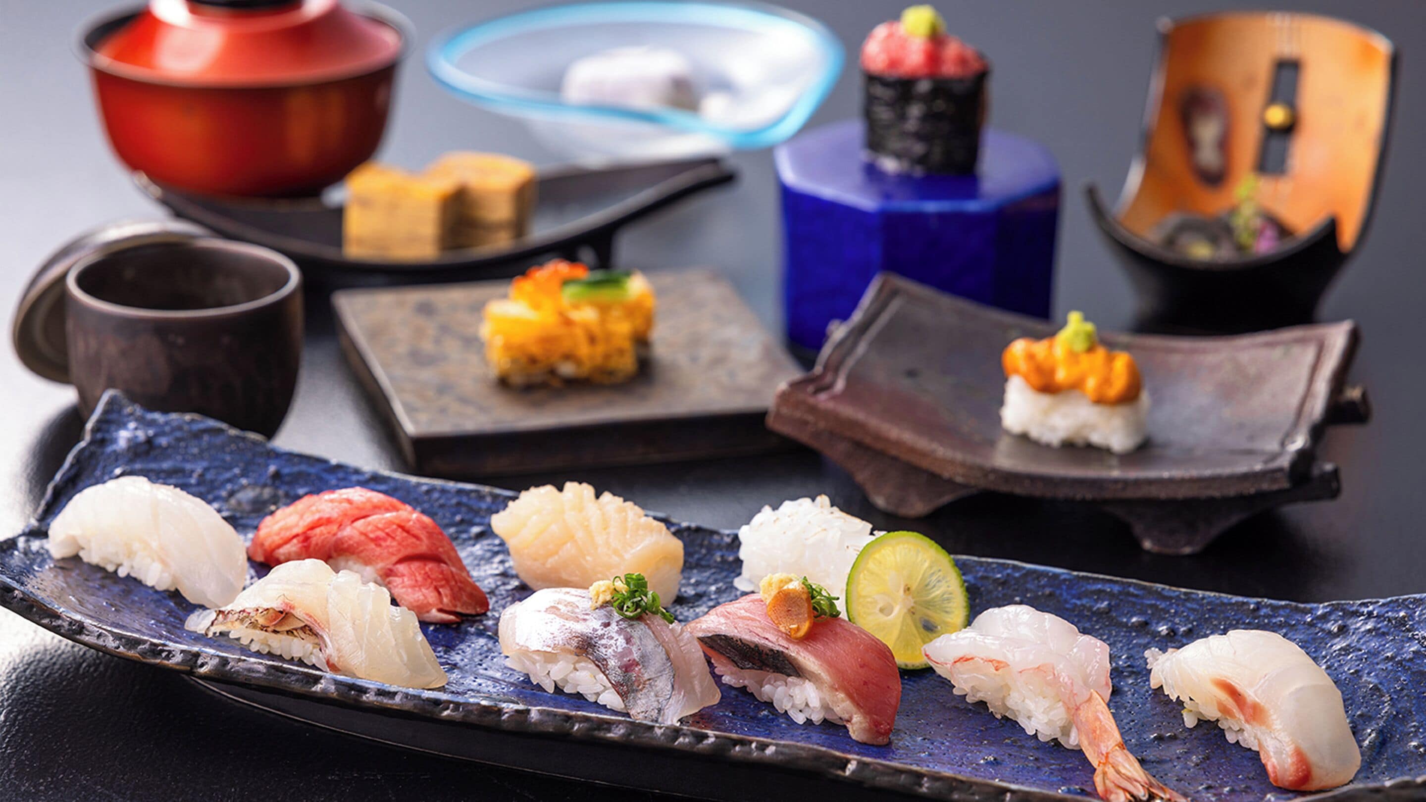 A variety of sushi dishes on a blue plate, with a small bowl and a cup in the background.