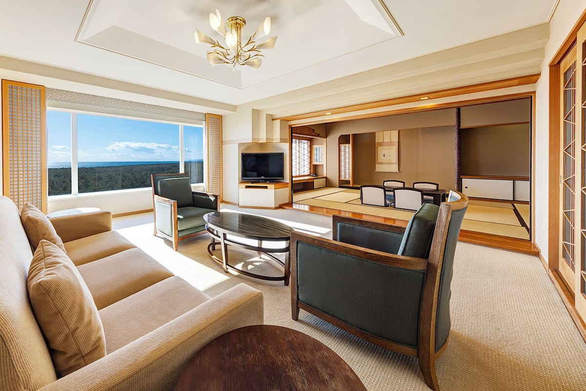 A modern living room with a sofa, a coffee table, and a TV. There is a dining area with a table and chairs. The room has large windows with a view of the outdoors.