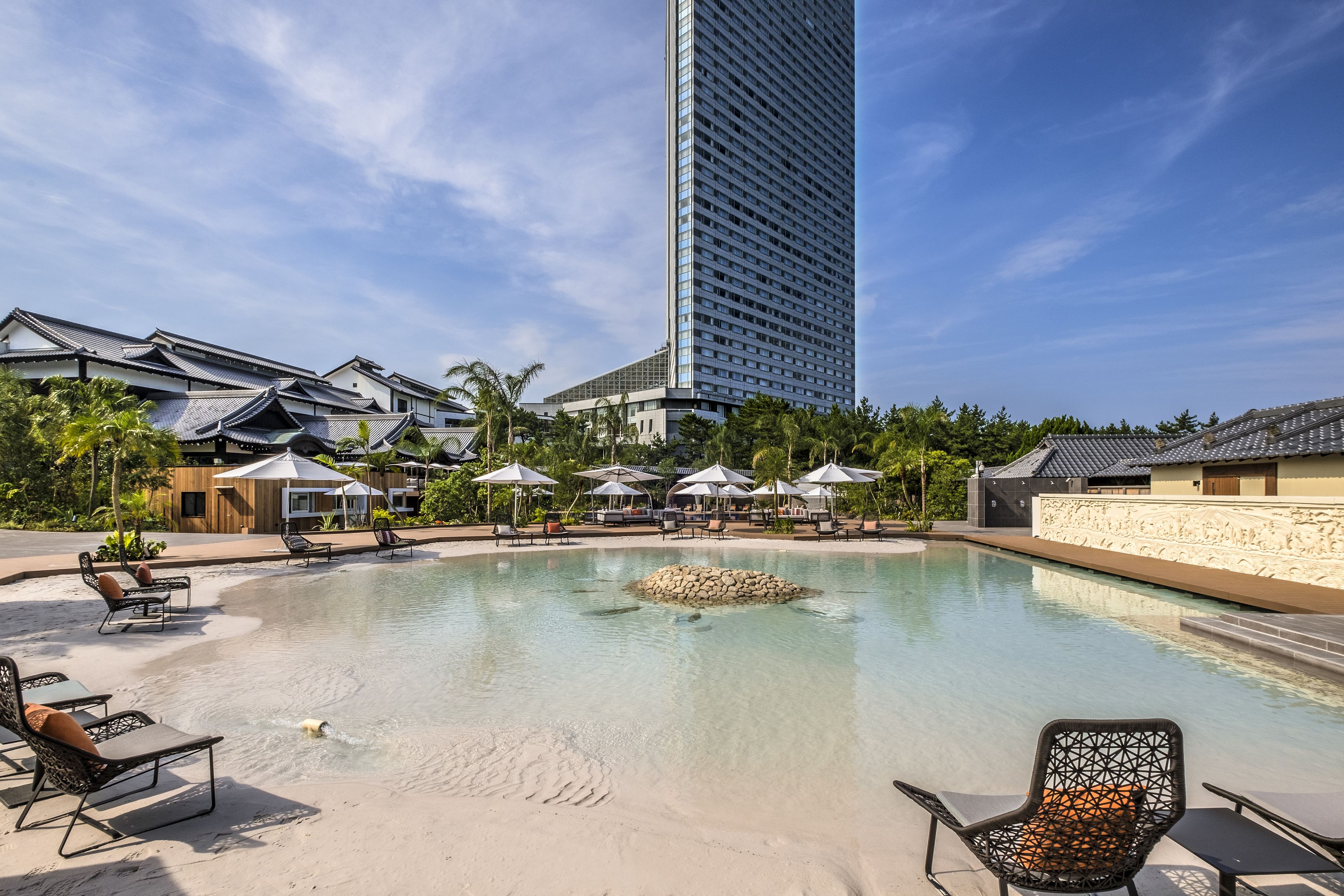 A pool area with lounge chairs, a stone centerpiece, and traditional-style buildings in the background.