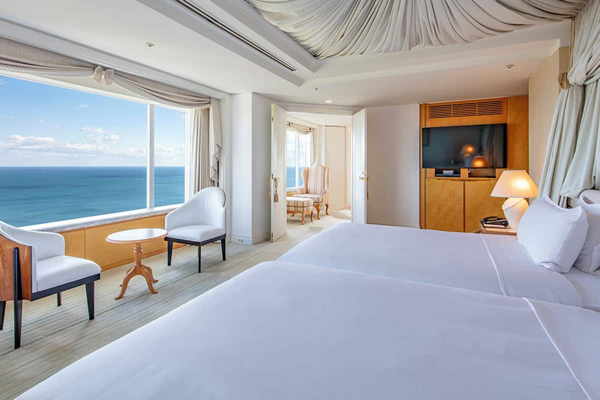 A hotel room with a large bed, two chairs, a small table, a TV, and a view of the ocean.