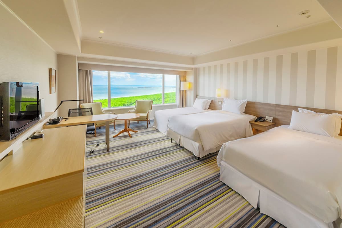 A hotel room with two beds, a desk, a TV, and a view of the ocean.