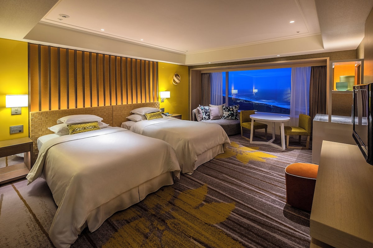 A hotel room with two beds, a small table, a sofa, and a view of the city at night.