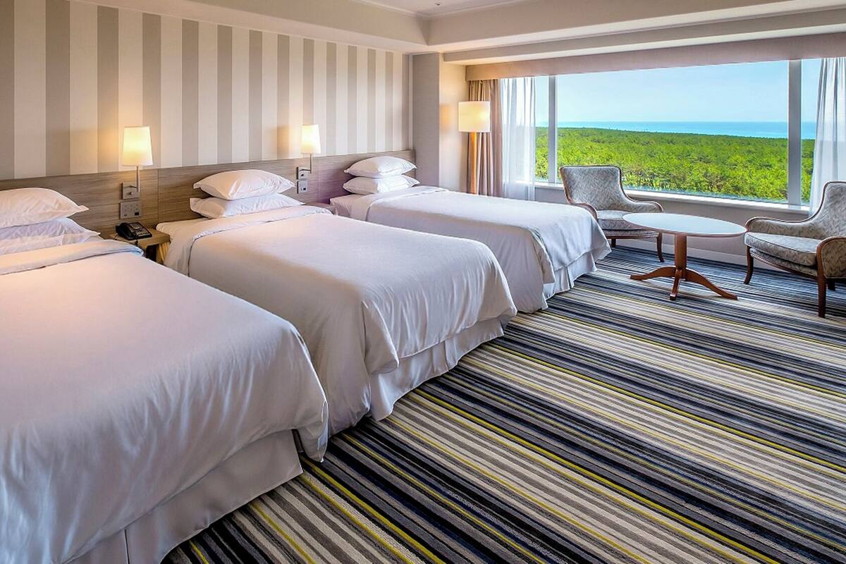 A hotel room with three beds, a striped carpet, a view of the ocean, and a small round table with a chair.