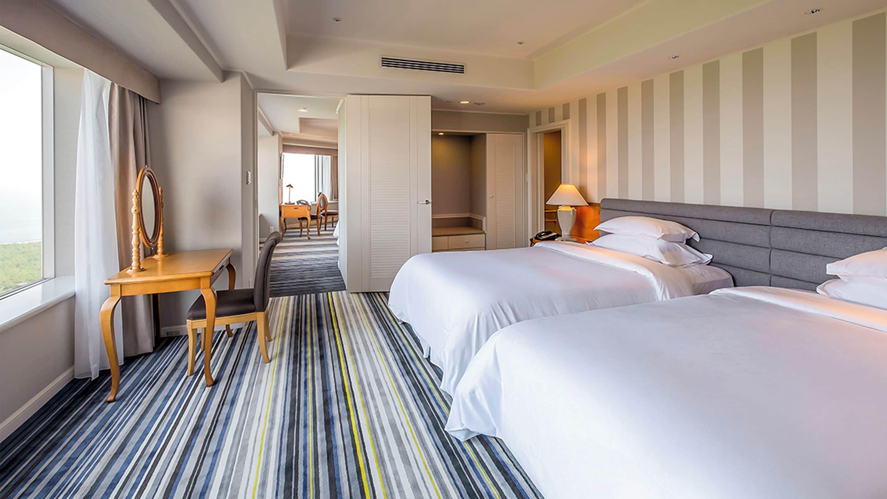 Habitación de hotel con dos camas, un escritorio, una silla, una lámpara y vistas a través de una puerta abierta.