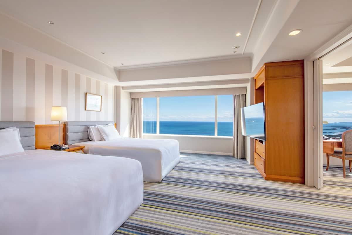 A hotel room with two beds, a TV, a desk, and a view of the ocean.