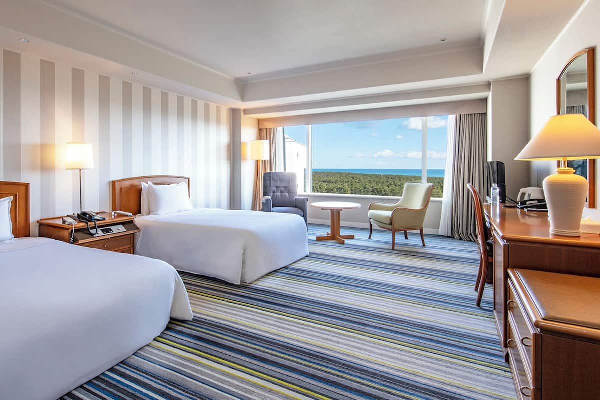 A hotel room with two beds, a small table, a chair, a desk, and a view of the ocean.