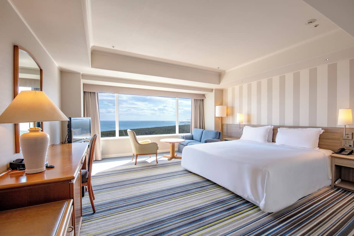 A hotel room with a large bed, a desk with a lamp, a chair, a small table, and a view of the ocean through large windows.