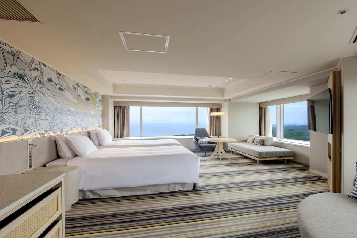 A hotel room with two beds, a sofa, a TV, and a large window with a view.