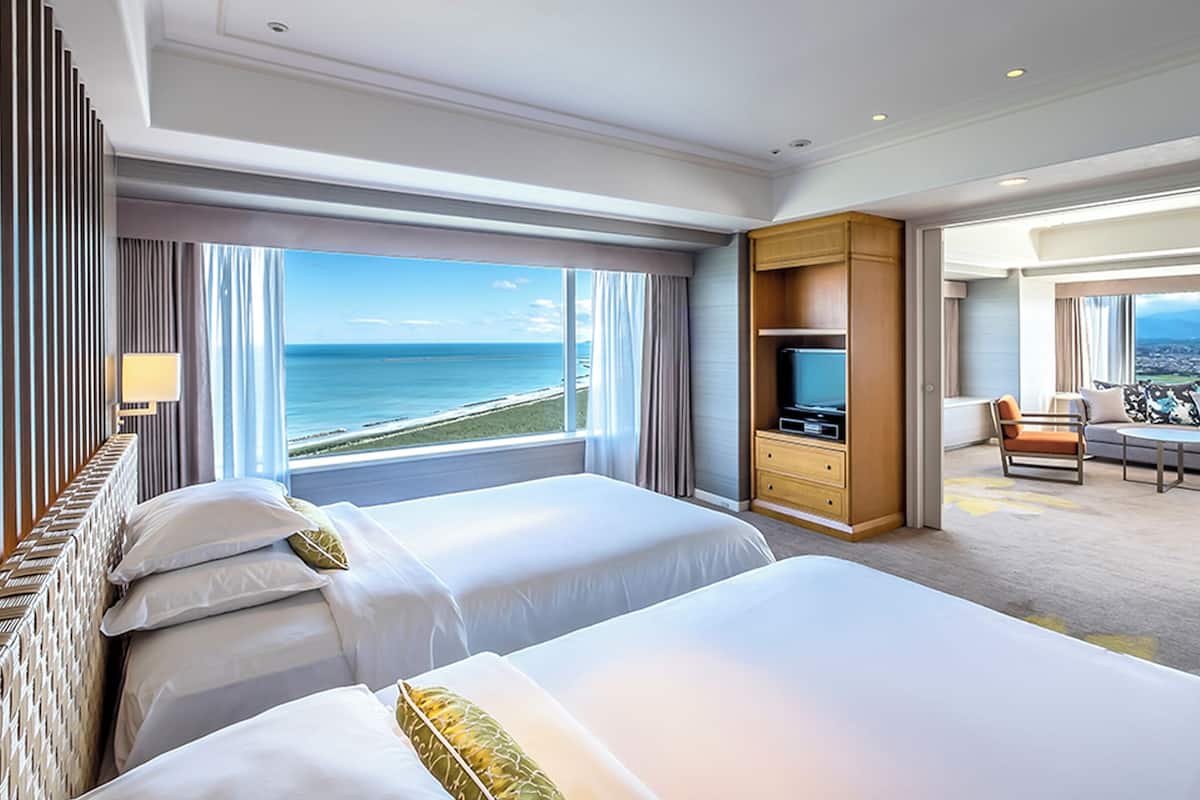 A hotel room with two beds, a TV, a desk, and a view of the ocean.