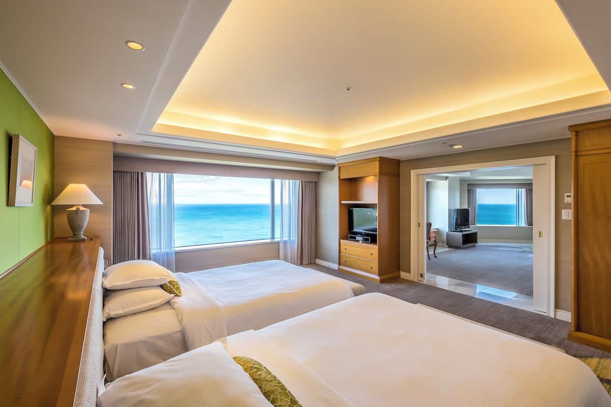 A hotel room with two beds, a TV, a desk, and a view of the ocean.