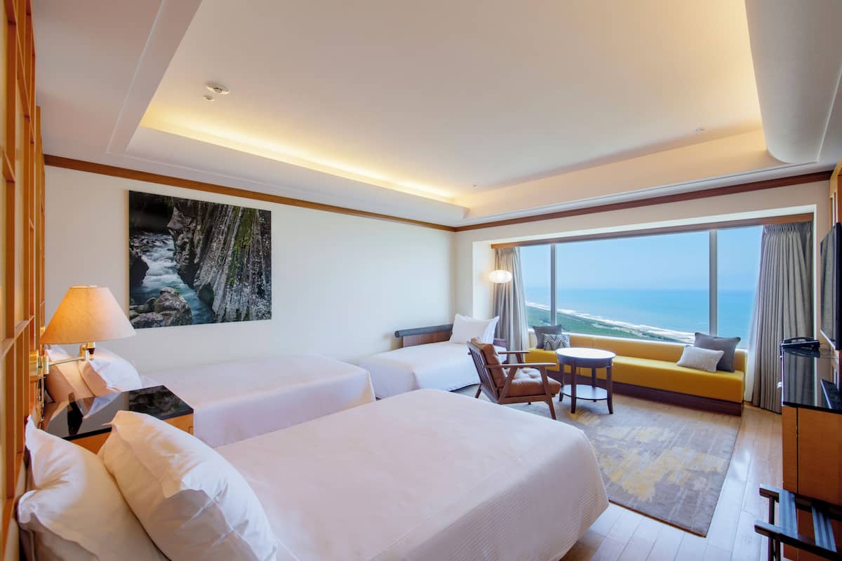 A hotel room with two beds, a sofa, a chair, a small table, a lamp, and a painting of a waterfall.