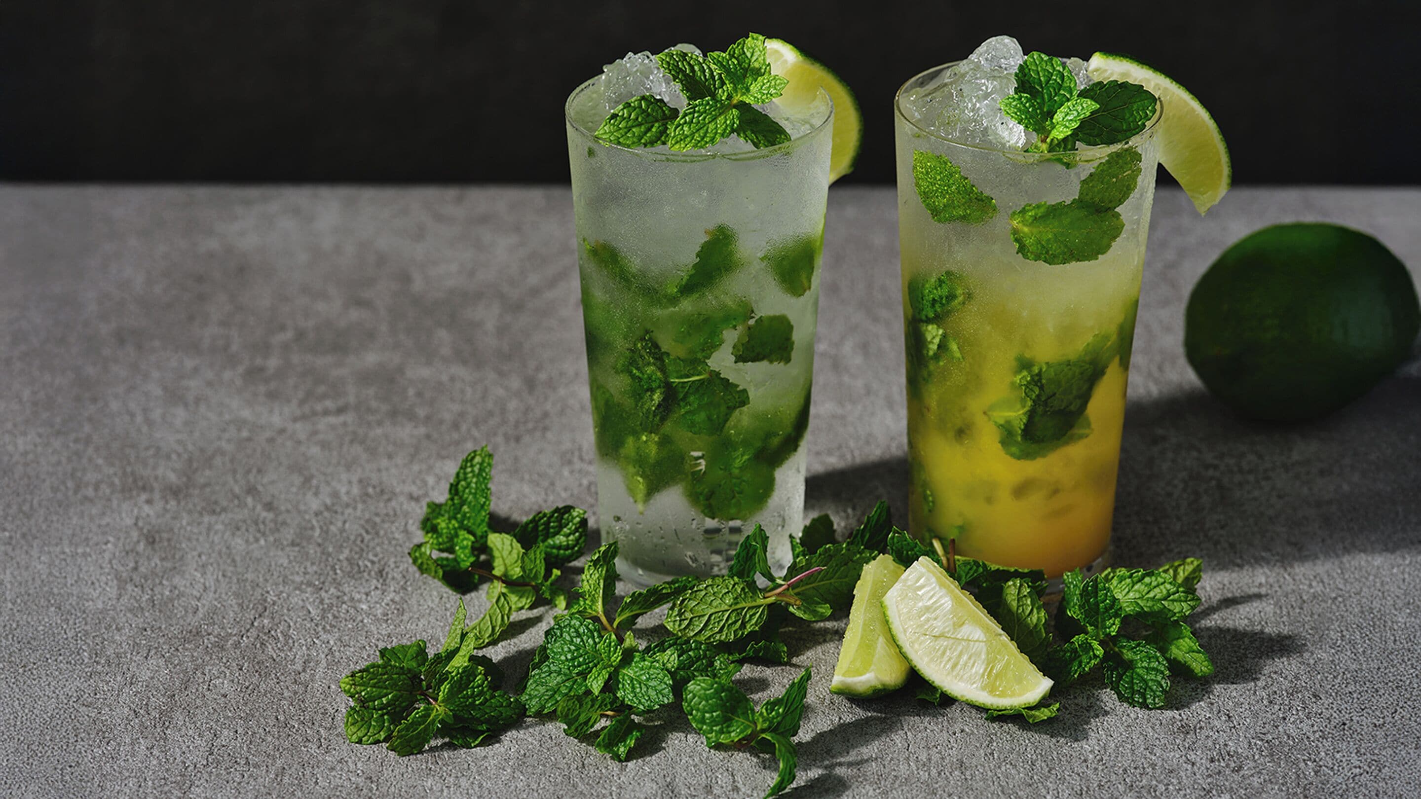 Two tall glasses of a green beverage with mint leaves and lime slices, ice cubes, and a lime wedge on the rim.